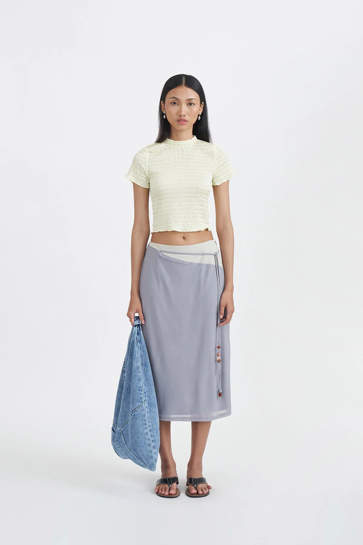 Woman wearing a light-yellow top by Callie Cotton and gray skirt holding a denim jacket on a white background