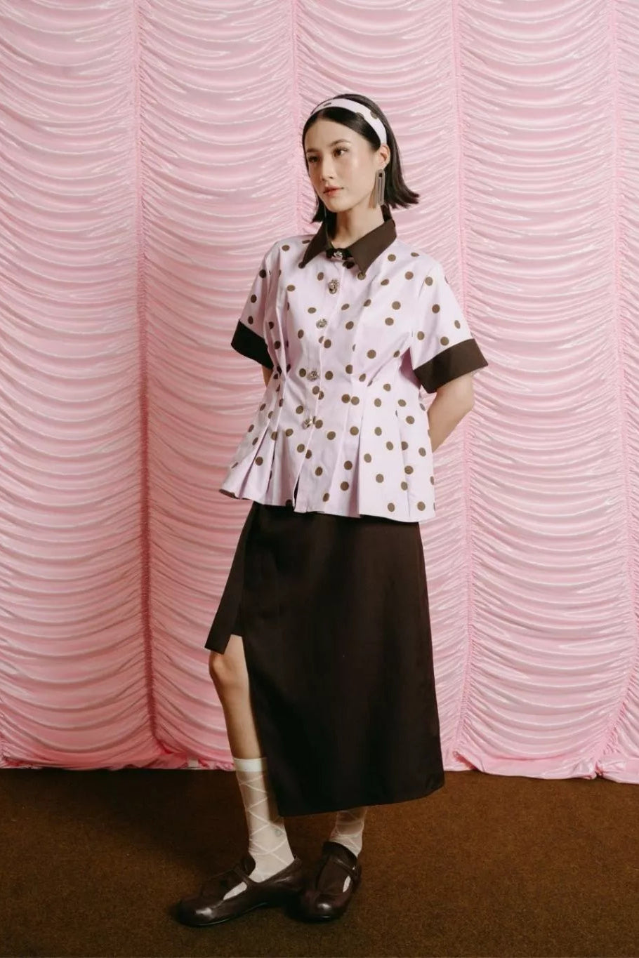 Woman wearing a polka dot blouse by LR Studio and black skirt against a pink textured wall.
