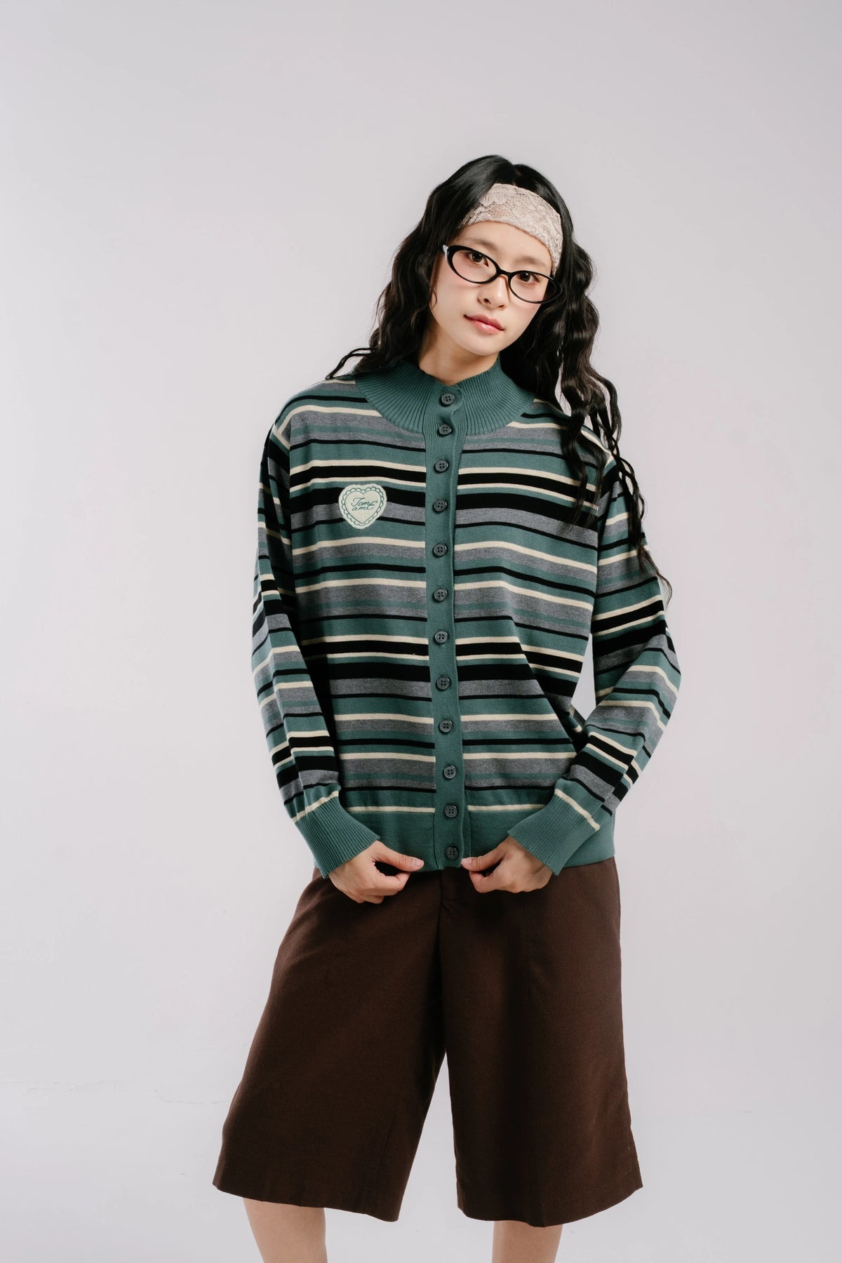 A model wearing a green and black striped button-up cardigan by Tome Ame with a small shield patch on the chest, paired with brown shorts, against a light gray studio background.