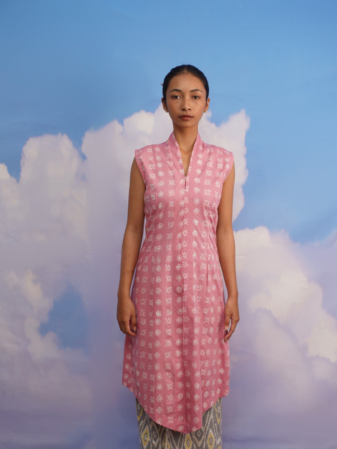 Woman wearing a pink sleeveless dress by Manikan with a pattern against a blue sky and cloud background