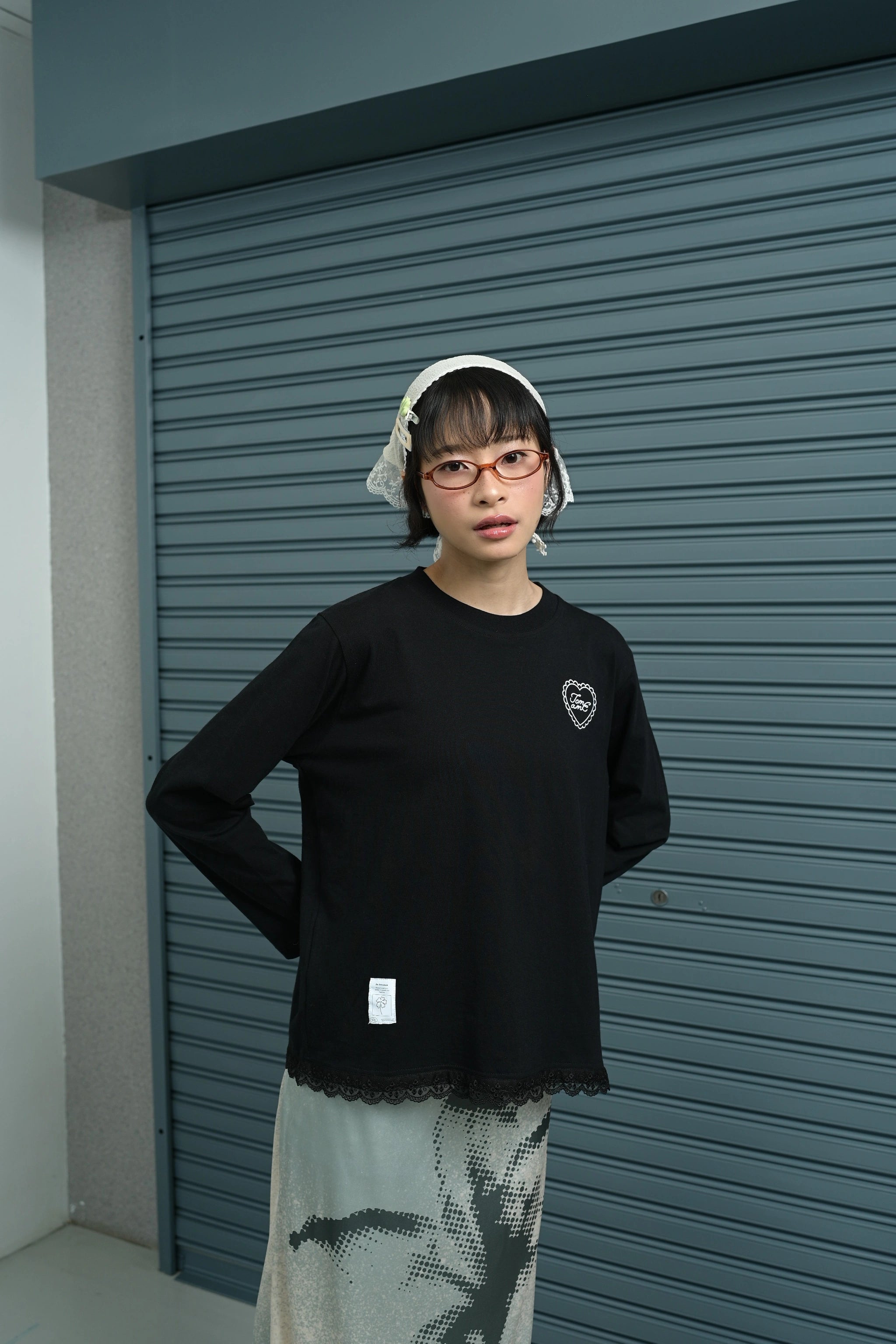 Person wearing a black long-sleeve t-shirt by Tome Ame with a heart design and lace details in front of a gray shutter.