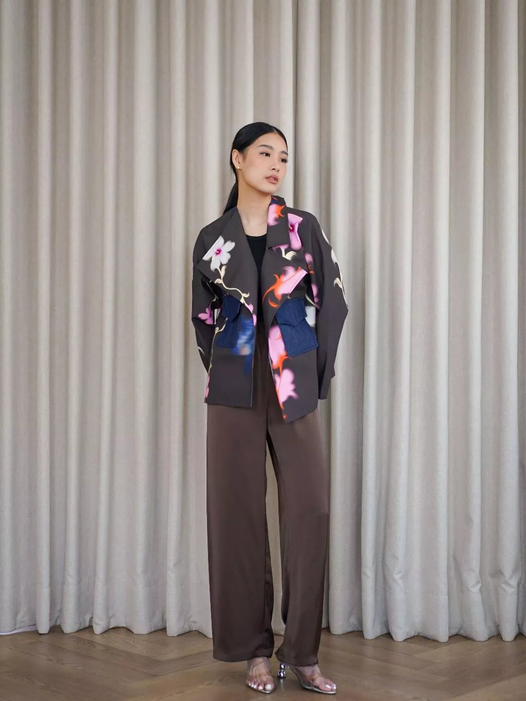 Woman wearing a colorful floral by LR Studio jacket and brown pants against a beige curtain background