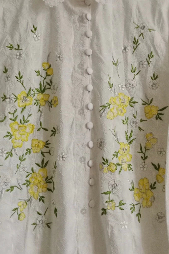 Close-up of a white or off-white embroidered shirt by Toko Didiyo with a button front, featuring yellow floral embroidery with green leaves and small white floral motifs.