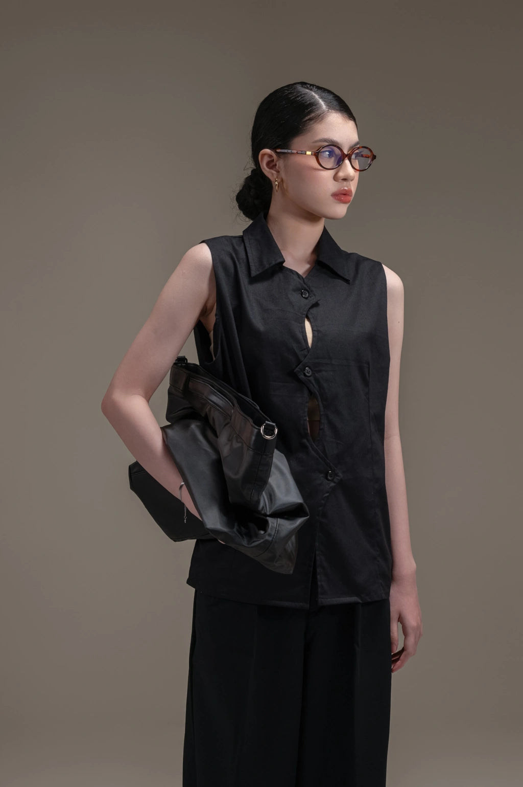 Woman wearing a black sleeveless blouse with cut-out design by Kajuaruna and holding a black bag against a brown background