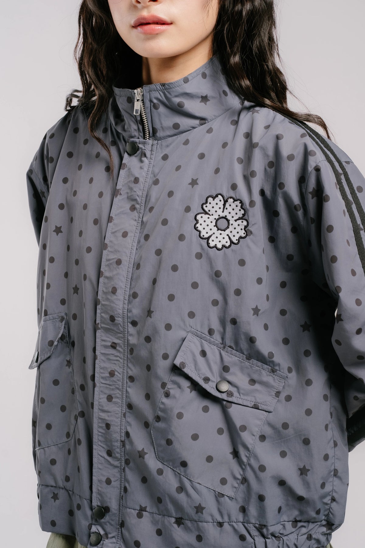 A close-up of a model wearing a gray zip-up jacket by Tome Ame with a black polka dot pattern, featuring a white and black flower applique on the chest.