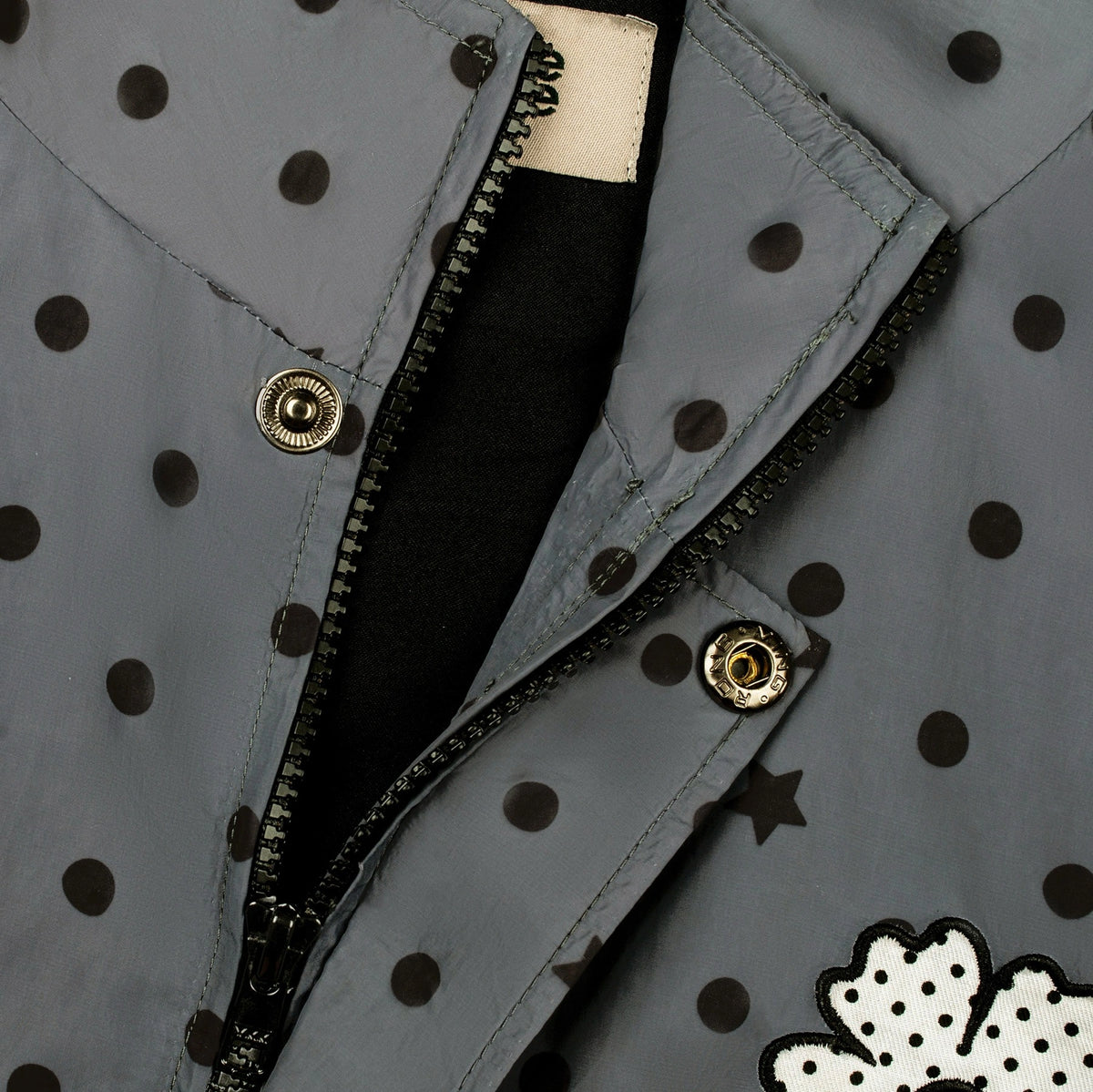 Close-up of a gray jacket with black polka dots, a black zipper, snap buttons, and a white applique patch with black polka dots in the bottom right corner.