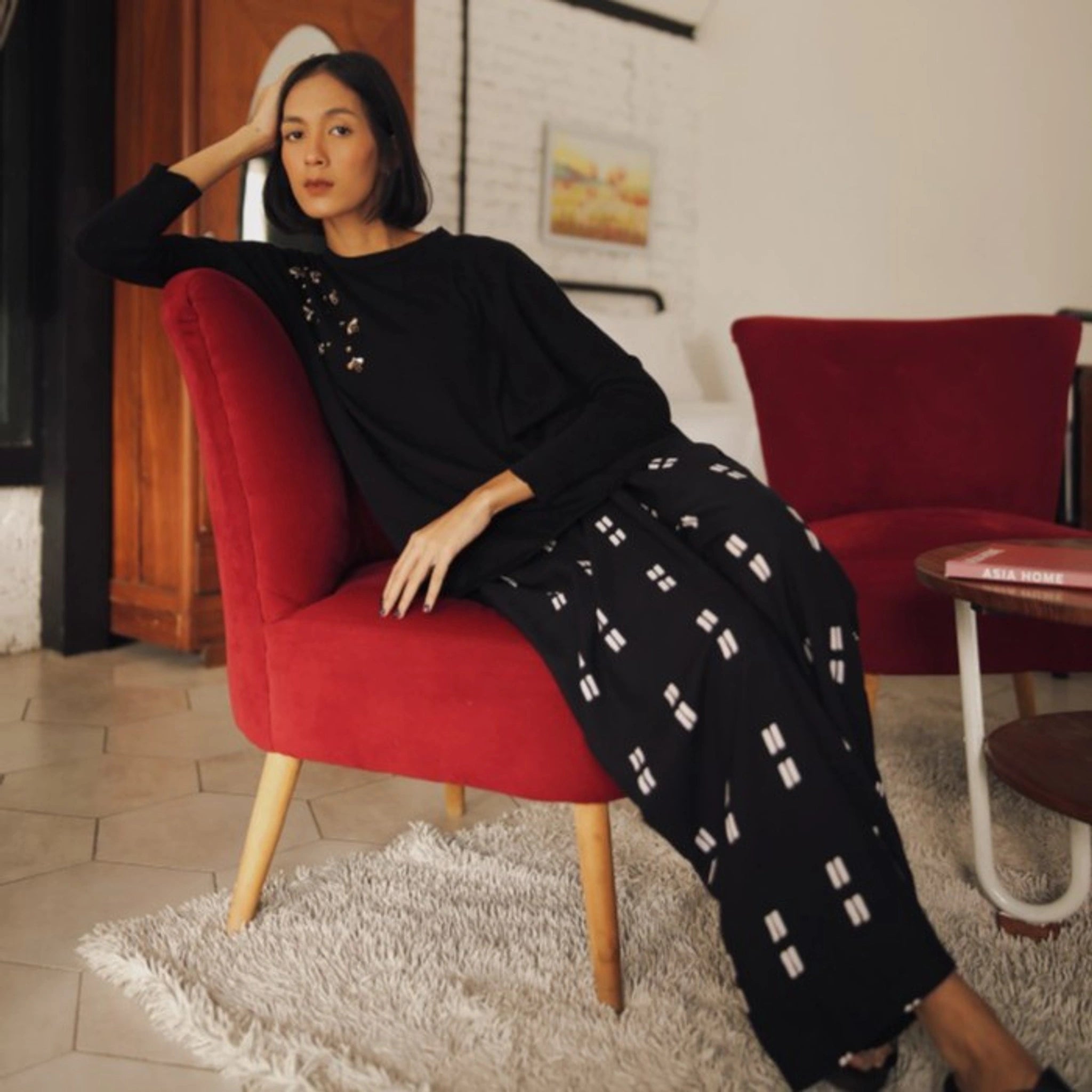 Woman in black beaded sweater from Damakara sitting on a red chair in a cozy room.
