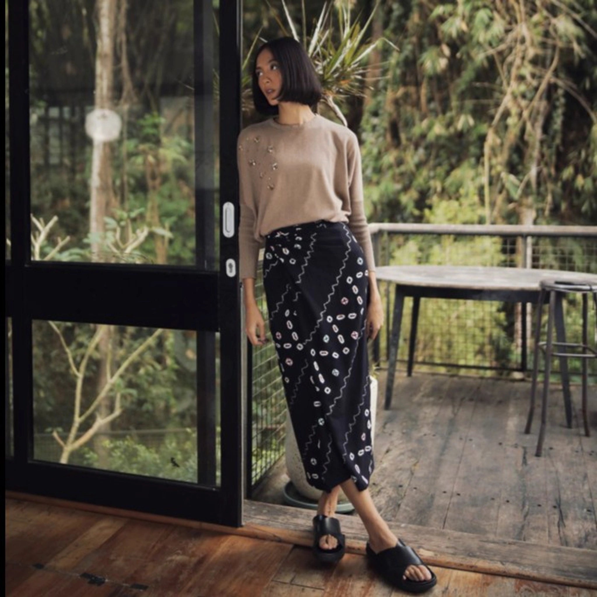 Woman standing in a room with a large glass door, wearing a light brown beaded sweater and black patterned skirt from Damakara.