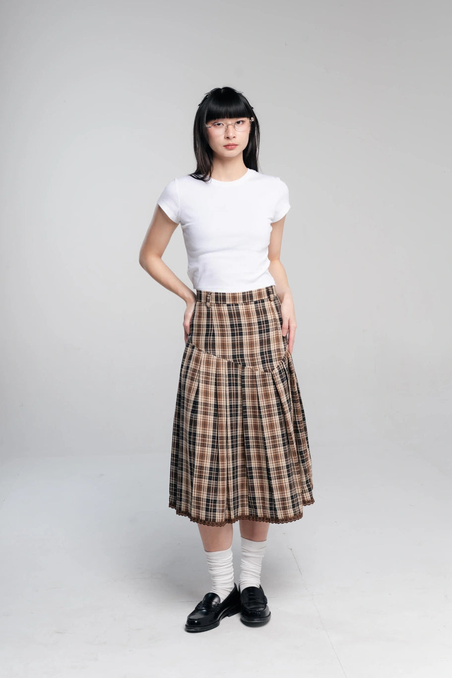 A model stands against a light gray studio background wearing a white short-sleeve crewneck t-shirt, a brown and beige plaid pleated midi skirt by Tome Ame, white socks, and black loafers.