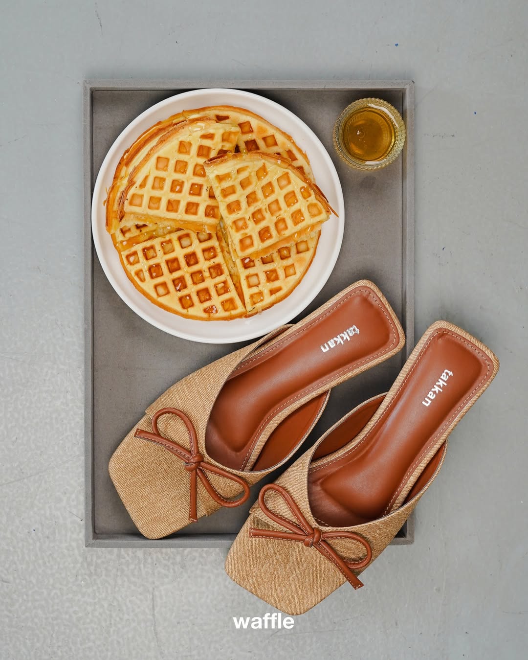 Pair of brown mules sandal by Takkan with a bow next to a plate of waffles on a gray surface.
