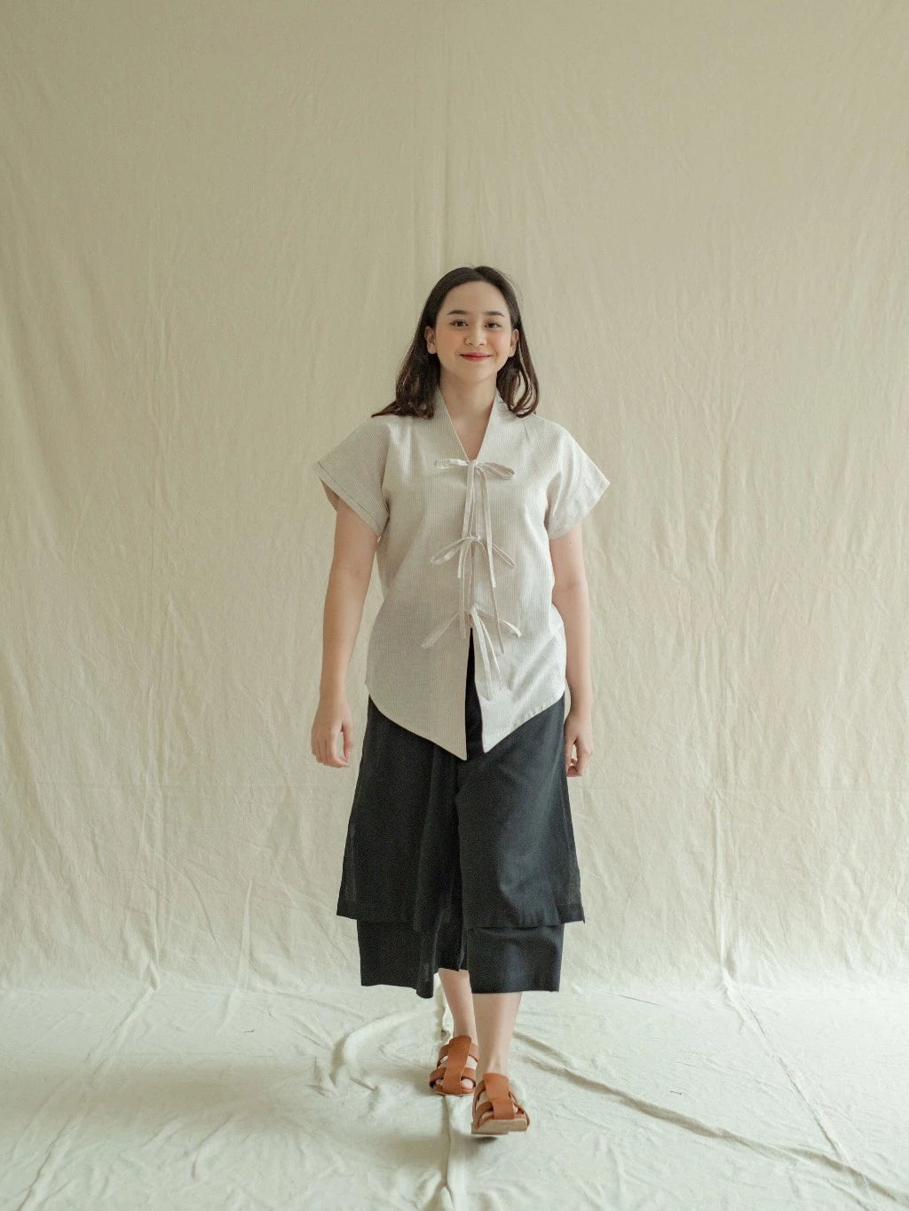 Woman wearing a cream short-sleeve kebaya and black pants against a plain background

Seorang wanita yang mengenakan kebaya lengan pendek berwarna krem dan celana hitam di depan latar belakang polos