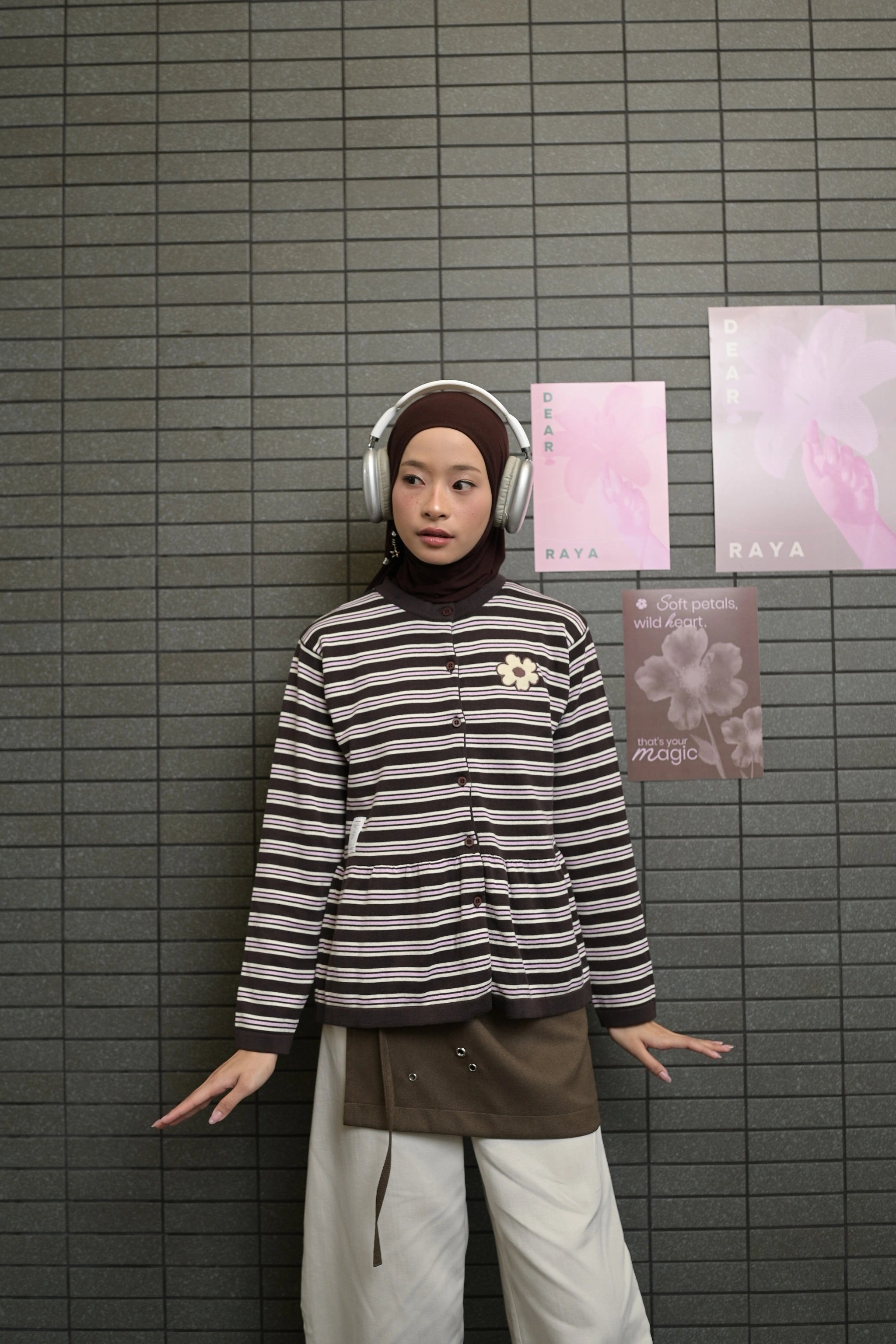 Person wearing a striped cardigans by Tome Ame and headphones against a tiled wall with posters.