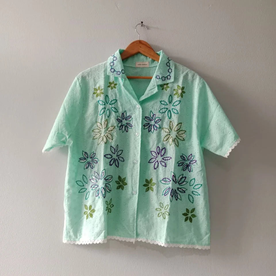 A mint green short-sleeve button-up shirt by Toko Didiyo with colorful floral embroidery, hanging on a wooden hanger against a plain light gray wall.
