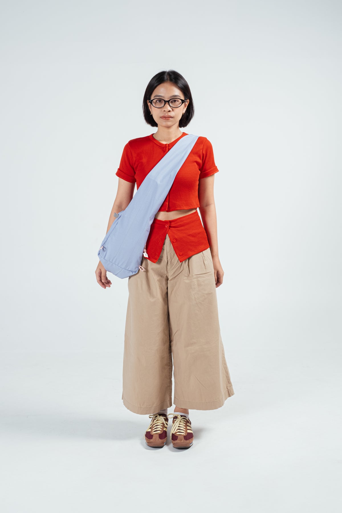 Person wearing a red top, beige pants, and a light blue crossbody bag by Measure Pleasure over one shoulder on a white background