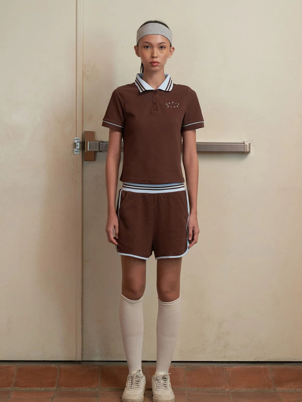 Person wearing a brown polo sports by Callie Cotton with a headband in front of a beige door.