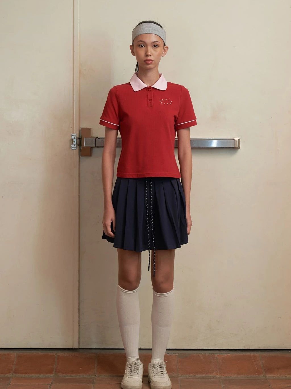Person wearing a red polo shirt by Callie Cotton with a white collar and a navy skirt against a plain wall.