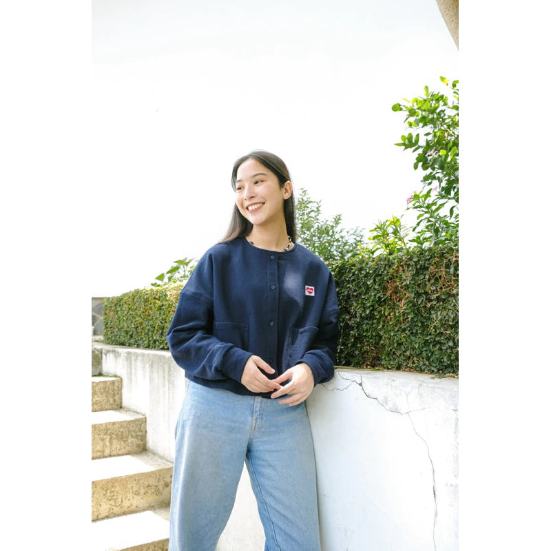 Person standing outdoors wearing a dark blue cardigan by Not Your Company and light blue jeans, leaning against a white wall with greenery in the background.