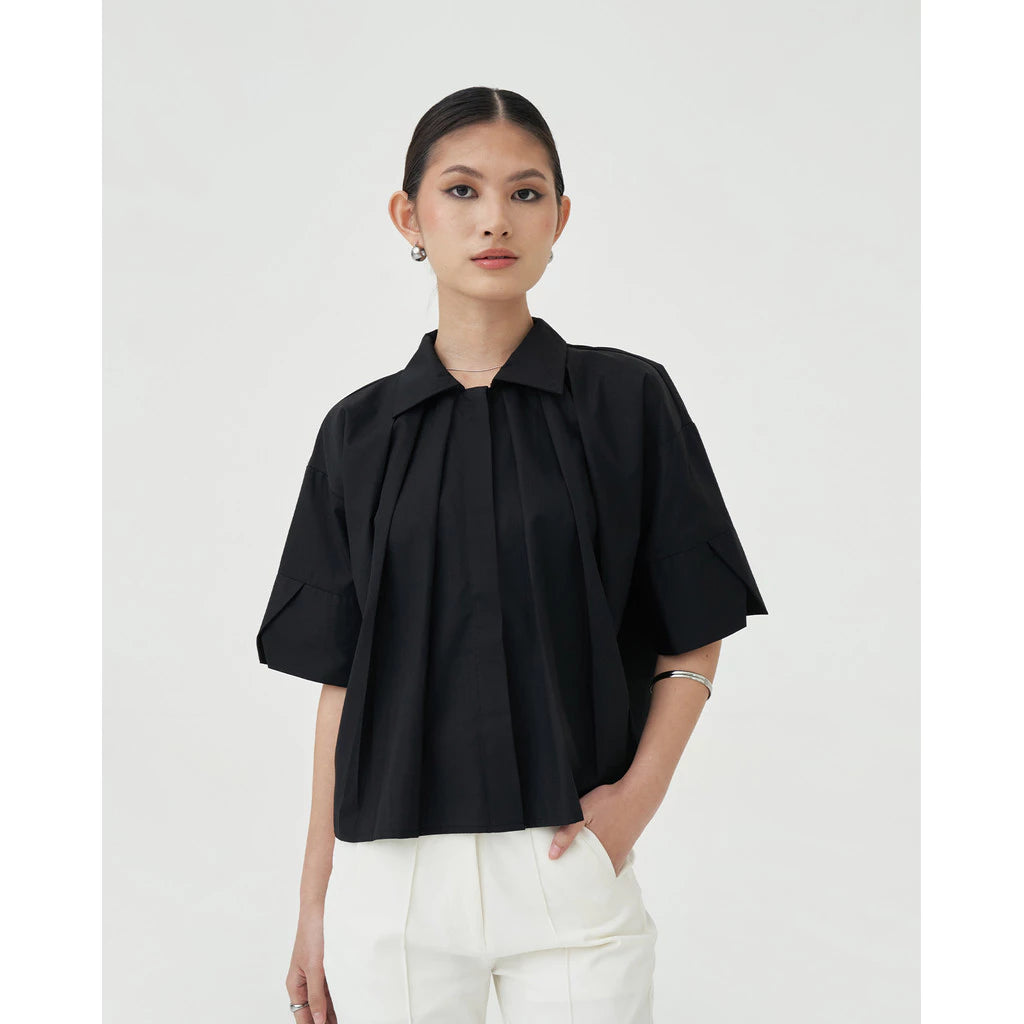 A woman wearing a black blouse by Achete de Nous with short sleeves and a round neckline, paired with white pants.