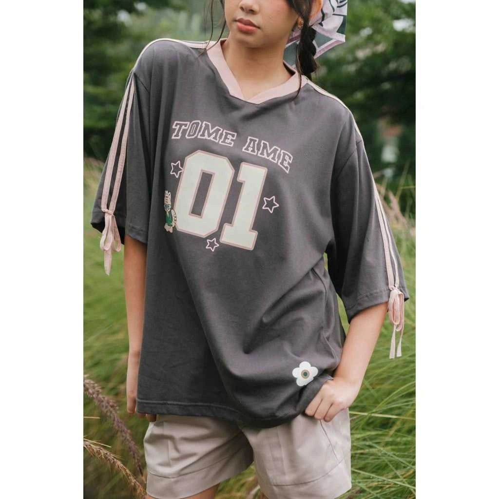 Person wearing a gray oversized t-shirt by Tome Ame with 'TOME AME 01' print in an outdoor setting.