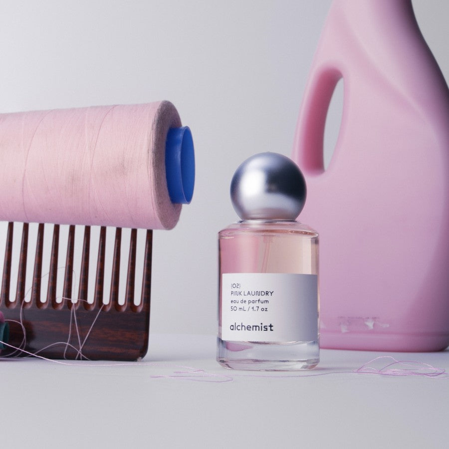 A pink bottle of Pink Laundry Alchemist fragrance next to a roll of pink fabric and a hairbrush, with a hint of a spilled thread and a clean, white round object in the background.