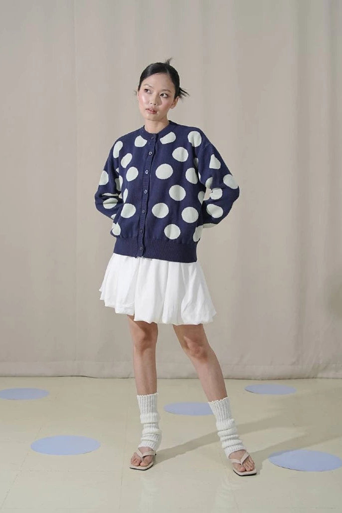 Person wearing a navy blue sweater with sage green polka dots by Tuff Puff and a white skirt on a beige background.