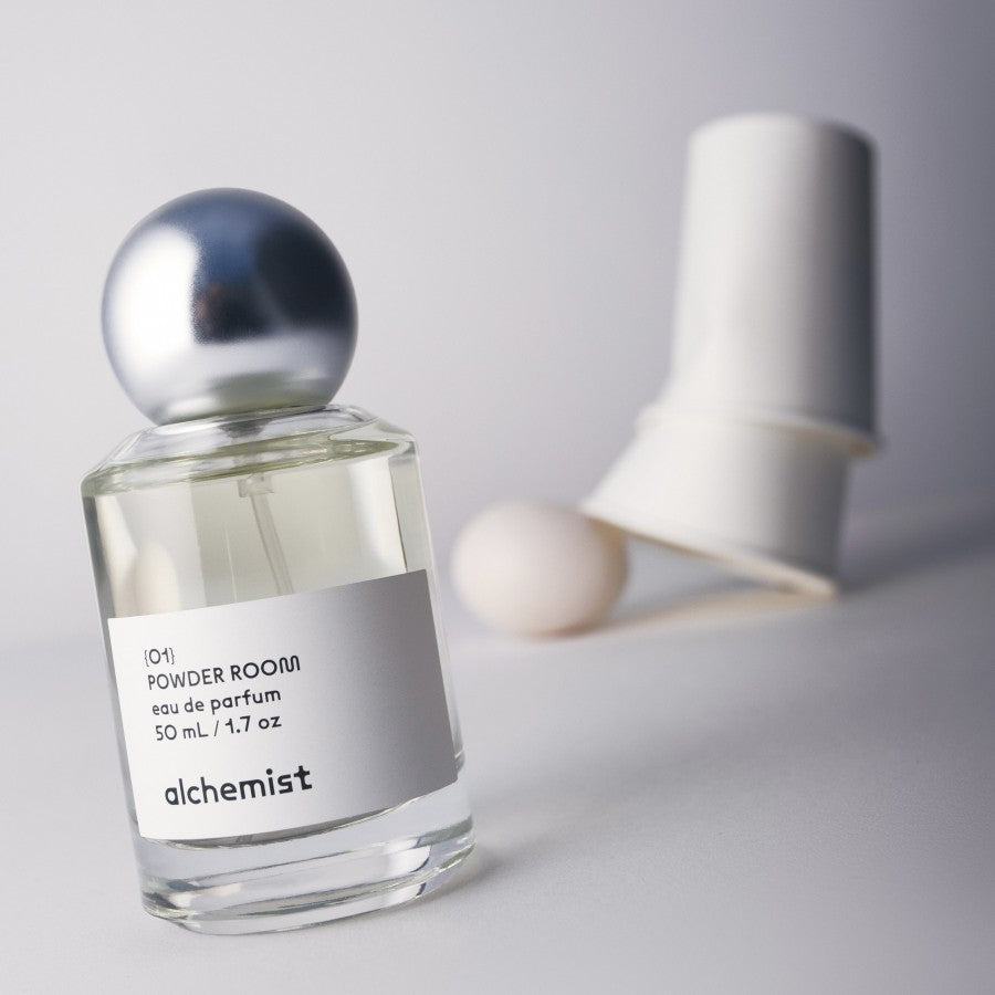 A bottle of 'Powder Room' Alchemist perfume with a metallic cap, displayed alongside a candle and a egg, on a white background.