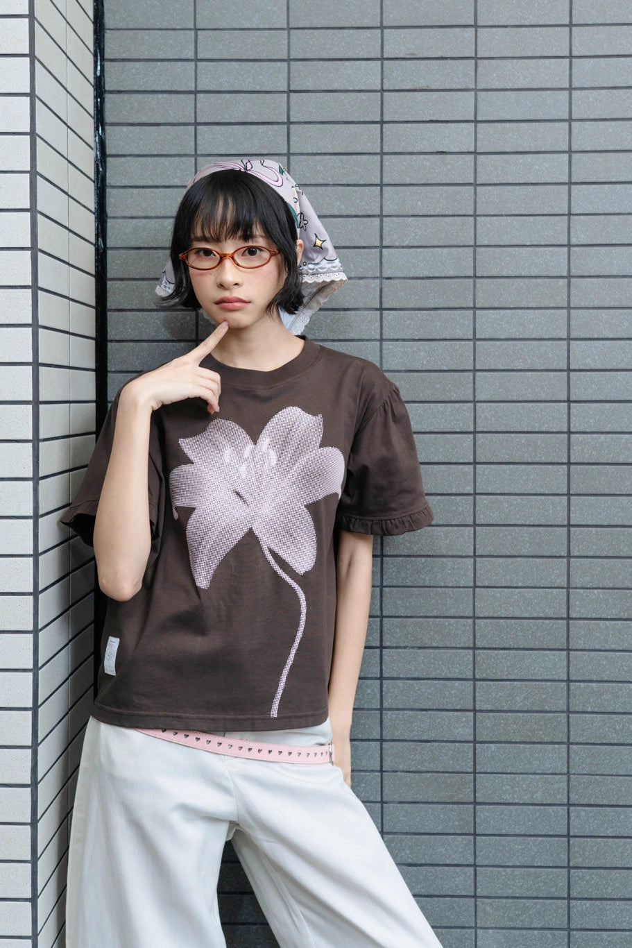A model wearing a dark brown t-shirt by Tome Ame with a large pale pink flower graphic, paired with white pants and a patterned headscarf, posing against a gray tiled wall.
