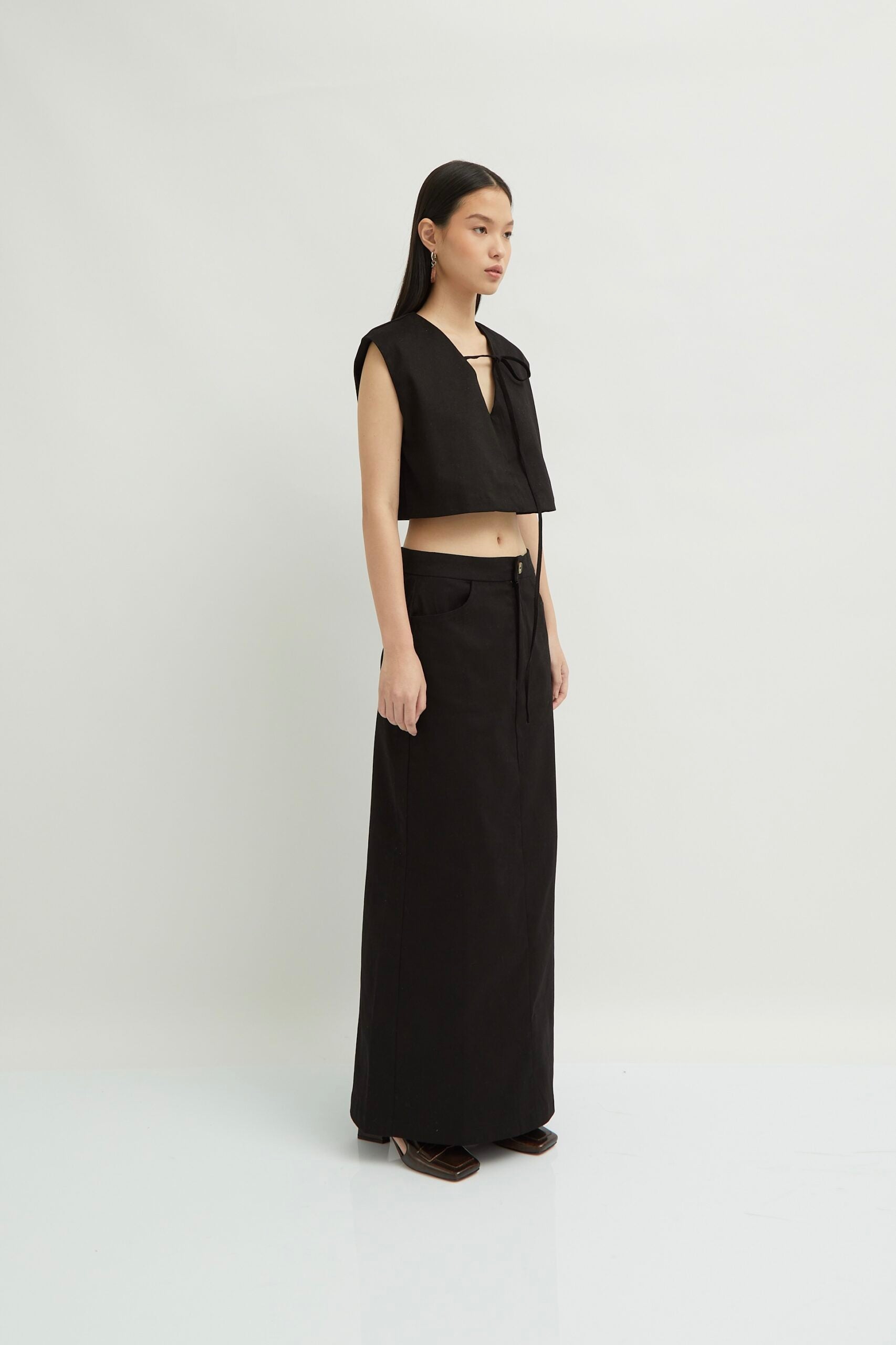 A model wearing a black maxi skirt with a front opening, paired with a black top and brown shoes, standing against a white background.