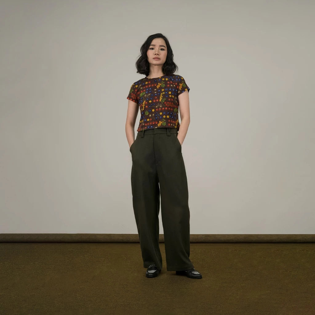 Woman wearing a patterned top and green barrel pants standing against a plain background
