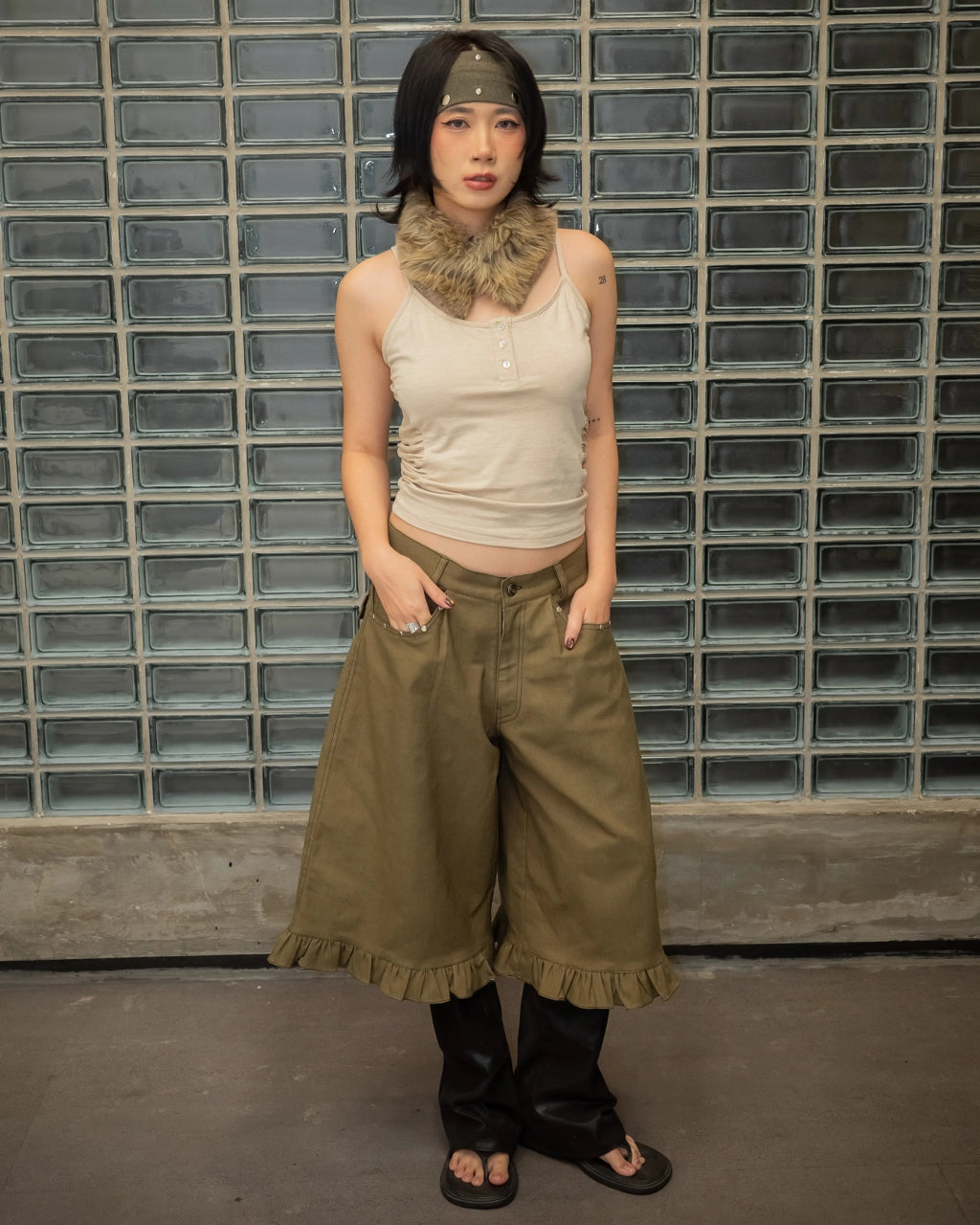 Woman wearing a beige sleeveless top and olive green wide-leg jorts pants against a glass block wall.
