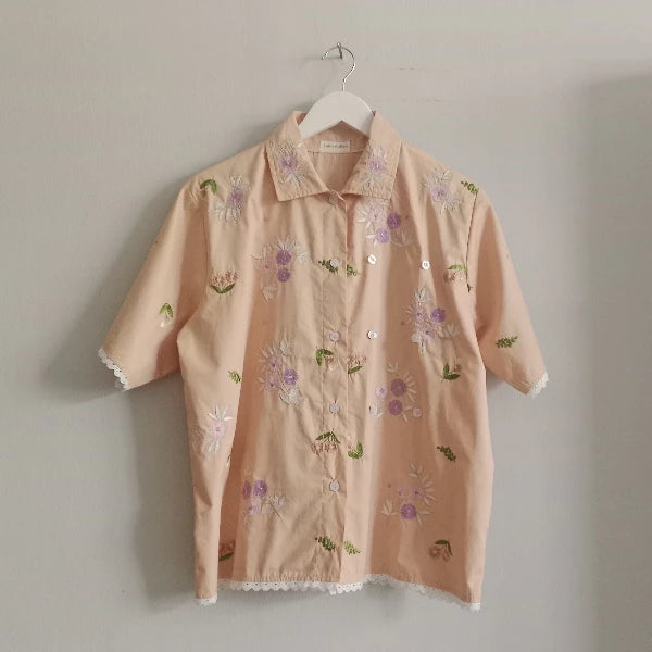 A short-sleeve button-up shirt by Toko Didiyo in a light peach-pink color, decorated with embroidered flowers in shades of purple, white, and green. The shirt features a pointed collar, white buttons, and white lace trim along the cuffs and hem, hanging on a white plastic hanger against a plain light gray wall.