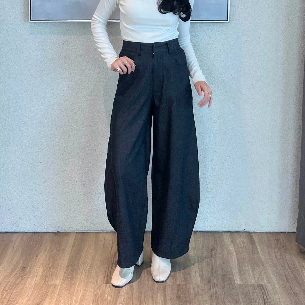 Person wearing a white long-sleeve top and black barrel pants standing against a light blue wall.