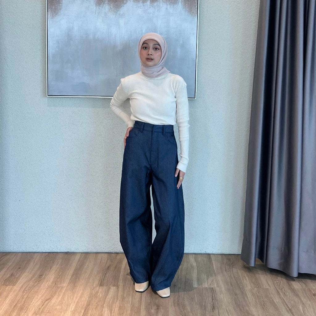 Person wearing a white sweater and navy barrel pants standing in a room with a neutral wall and curtain.