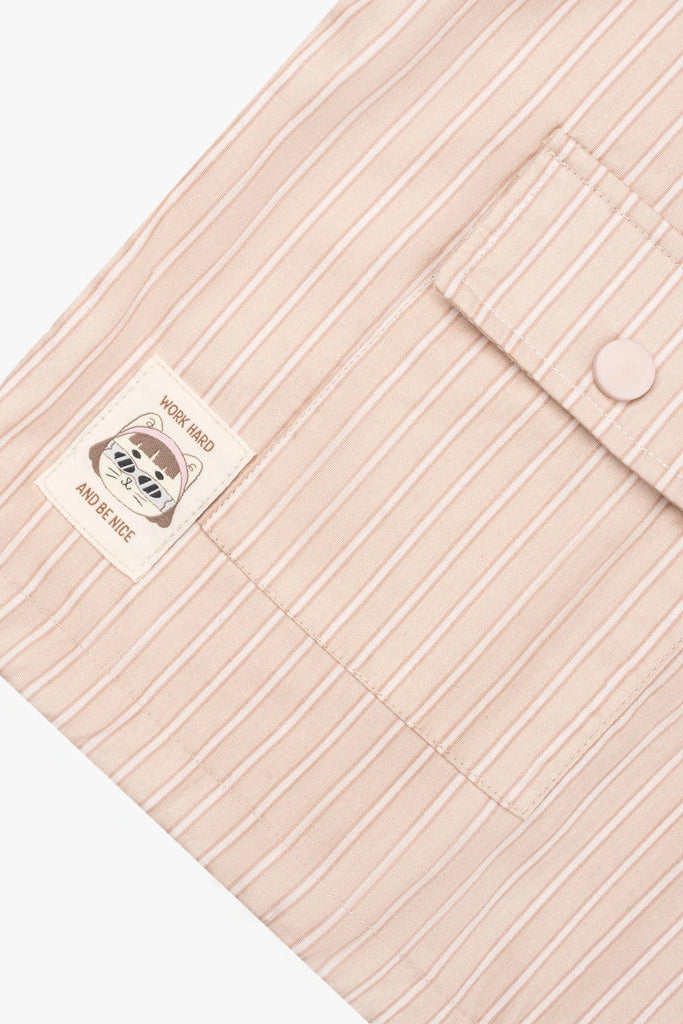 Close-up of a pale pink striped outer by Tome Ame with a buttoned pocket and a sewn-in label that reads 'WORK HARD AND STAY NICE' with a bear illustration.