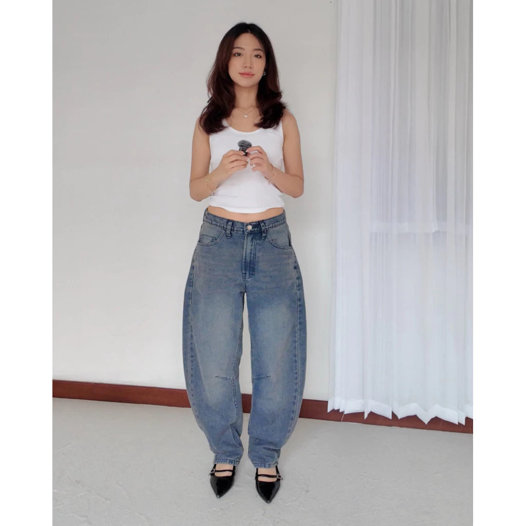 Woman wearing a white tank top and denim barrel pants standing against a plain white wall.