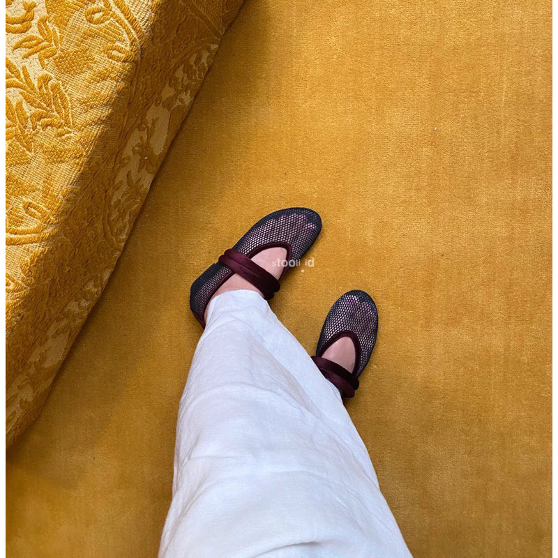 Person wearing black mesh flatshoes with maroon crisscross elastic strap by Stooli on a mustard yellow carpet