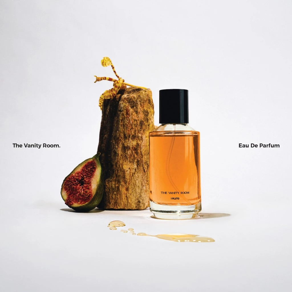 A bottle of 'The Vanity Room' perfume by Scents Of Pluto, accompanied by a piece of wood and a fig, against a white background.