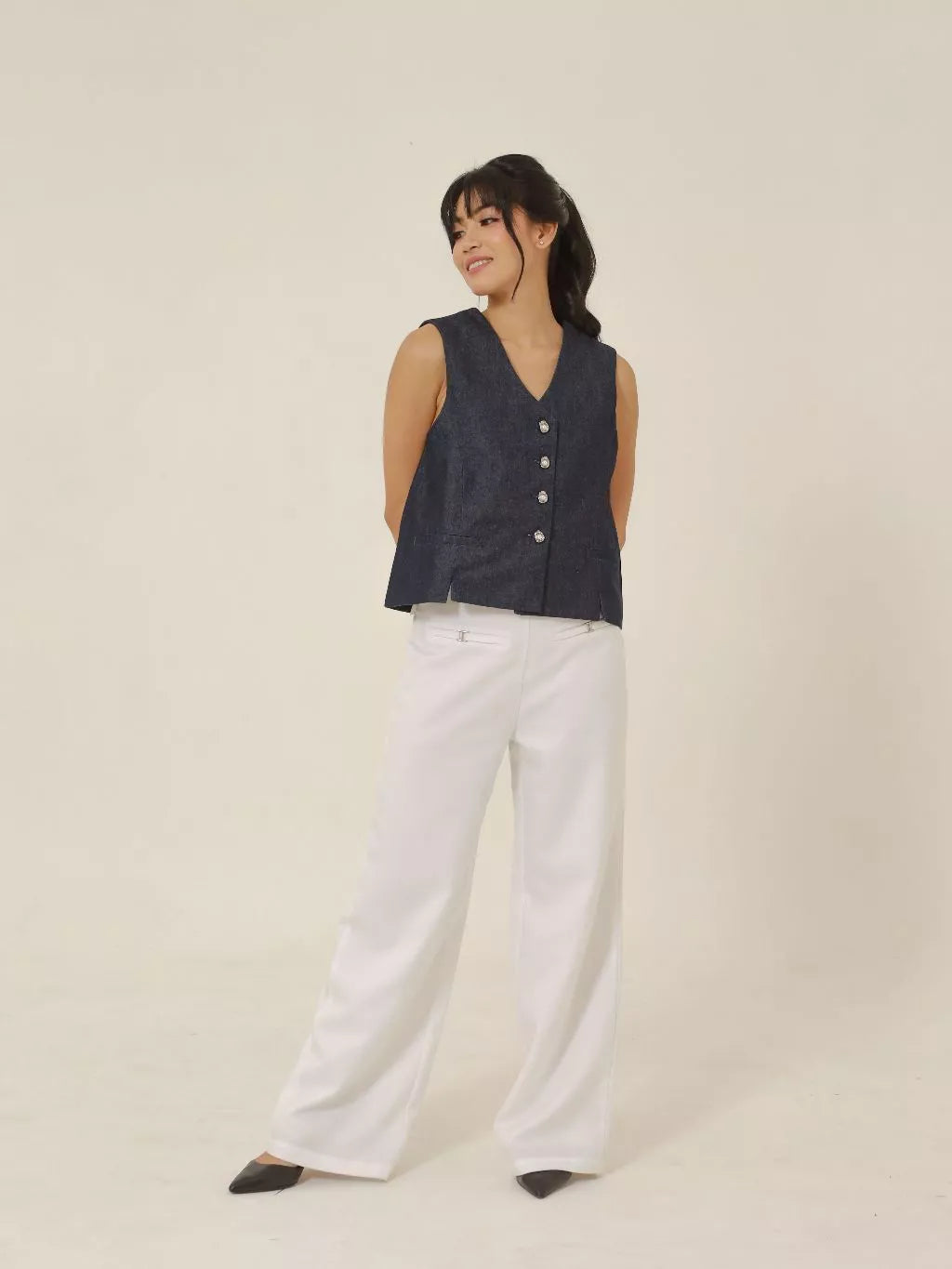 Woman wearing a dark denim vest over a white top and pants on a beige background