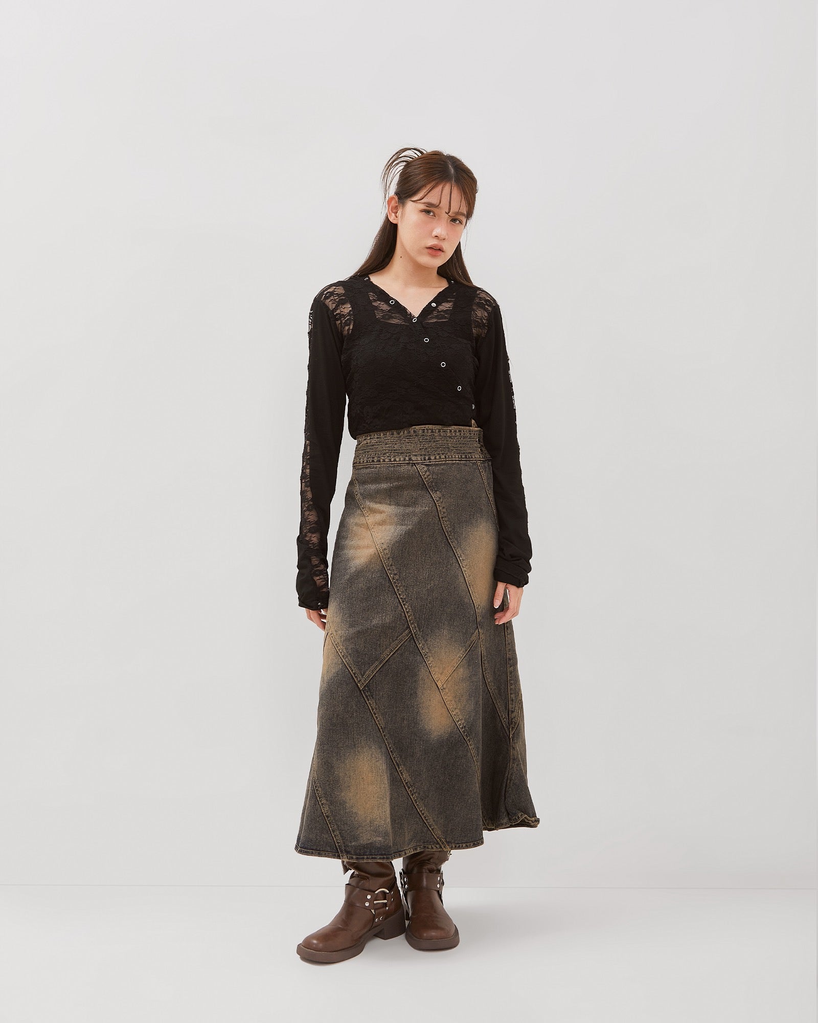A person standing wearing a long brown washed denim skirt with a flared silhouette, paired with a black top and boots, against a white background.