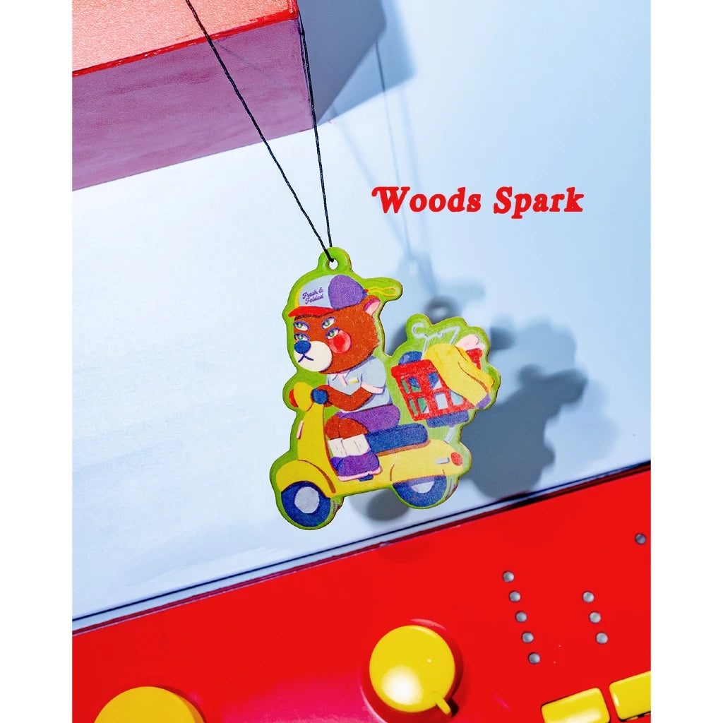 Colorful toy bear on a scooter air freshener by Oh.Irv with 'Woods Spark' branding against a red and blue background.