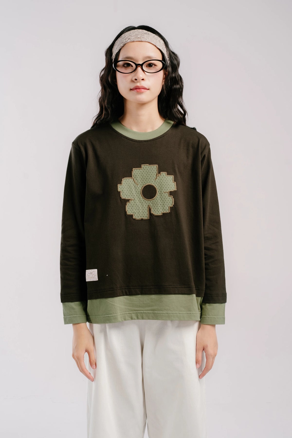 A young woman wearing a dark olive green long-sleeve t-shirt by Tome Ame with a textured light green flower graphic on the chest, paired with white pants and a beige headband, standing against a light gray studio background.