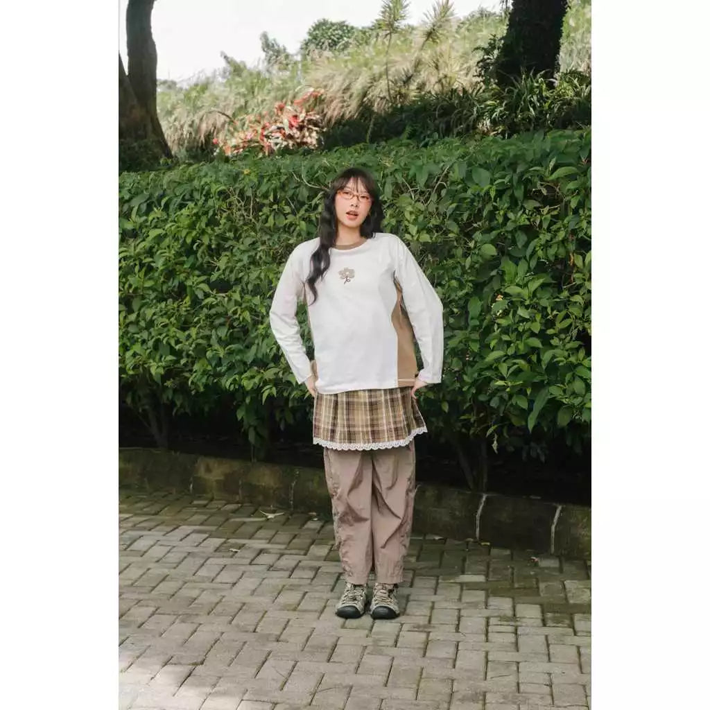 Person wearing a white long sleeve t-shirt by Tome Ame and plaid skirt standing in front of greenery.