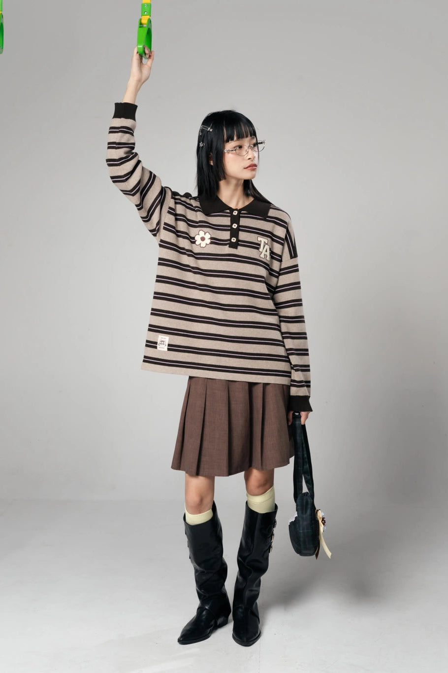 A model wearing a brown and beige striped long-sleeve polo shirt by Tome Ame, a brown pleated skirt, and black knee-high boots, holding a green bottle against a plain gray studio background.