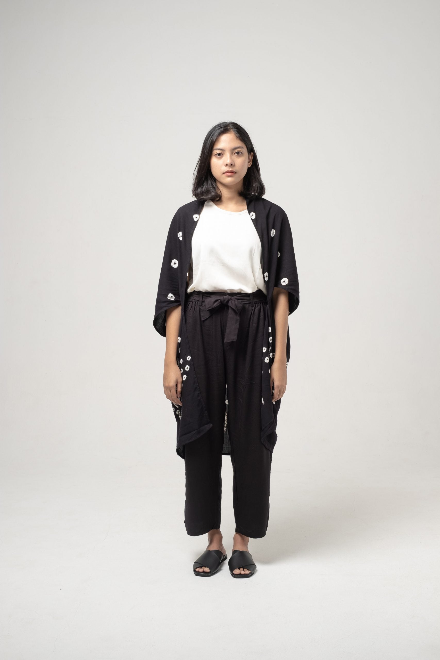 A woman standing against a white background wearing a black traditional Indonesian Seruni Outer with a patterned design from Damakara.